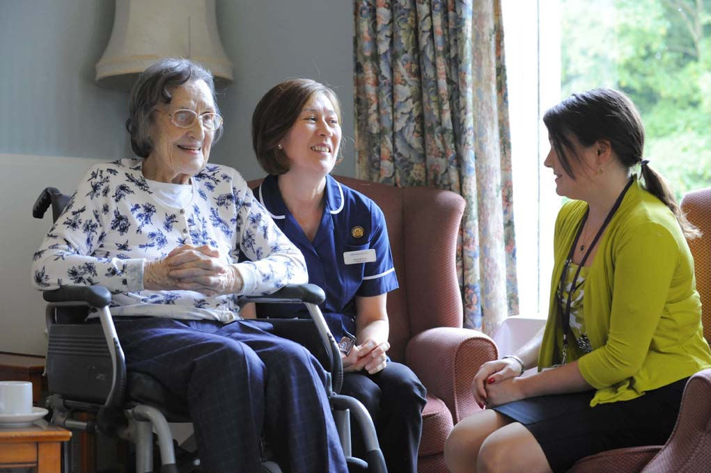 Image of an older white woman in a wheelchair being assisted in her home by 2 middle-aged white women health care workers.